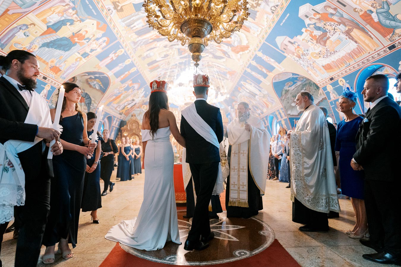 A couple standing in front of a priest during a traditional Eastern Orthodox wedding ceremony inside a beautifully decorated church, surrounded by family and friends.