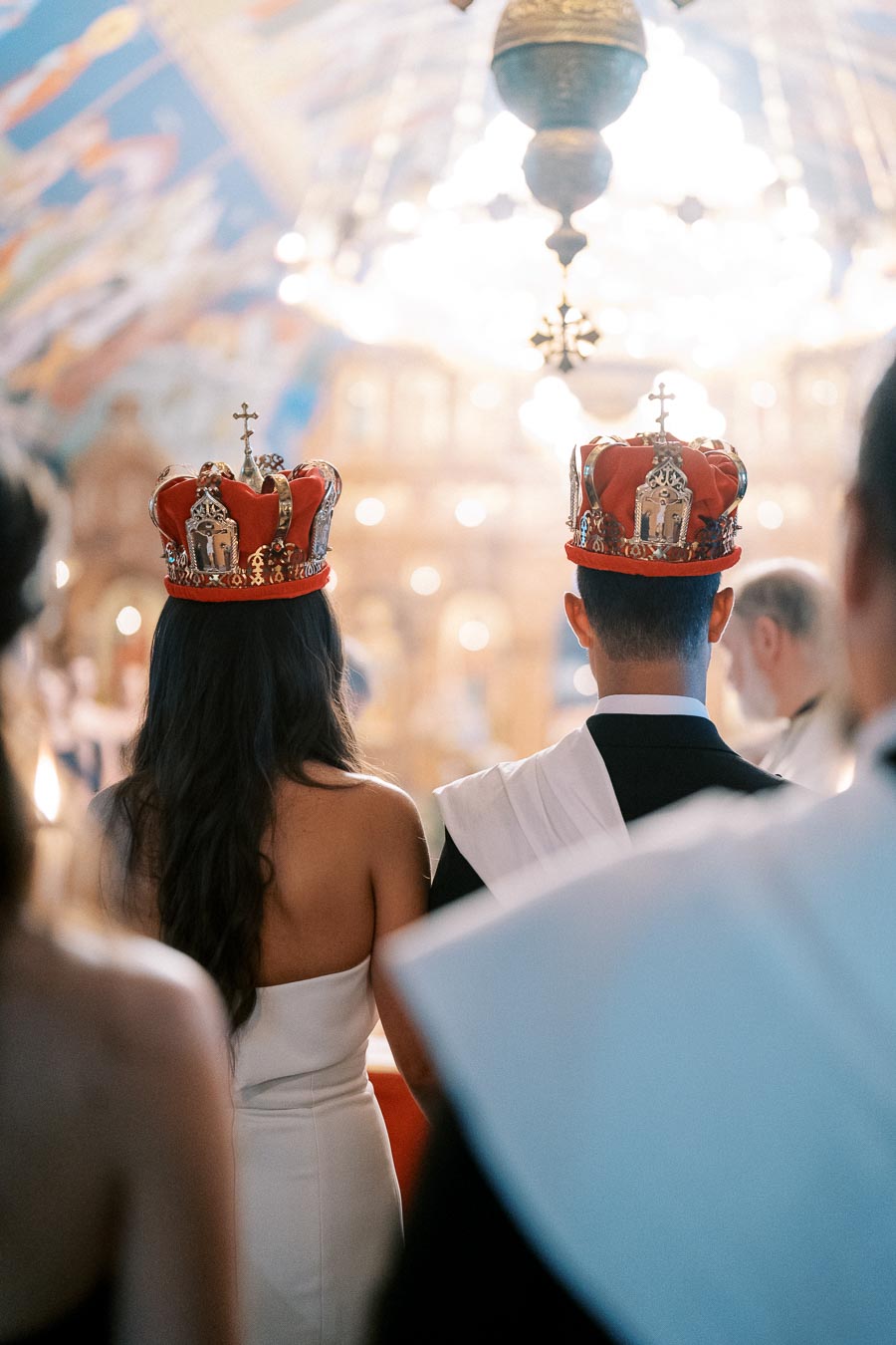 A bride and groom wearing ornate crowns during a traditional wedding ceremony in a richly decorated setting, with soft lighting and an elegant ambiance.