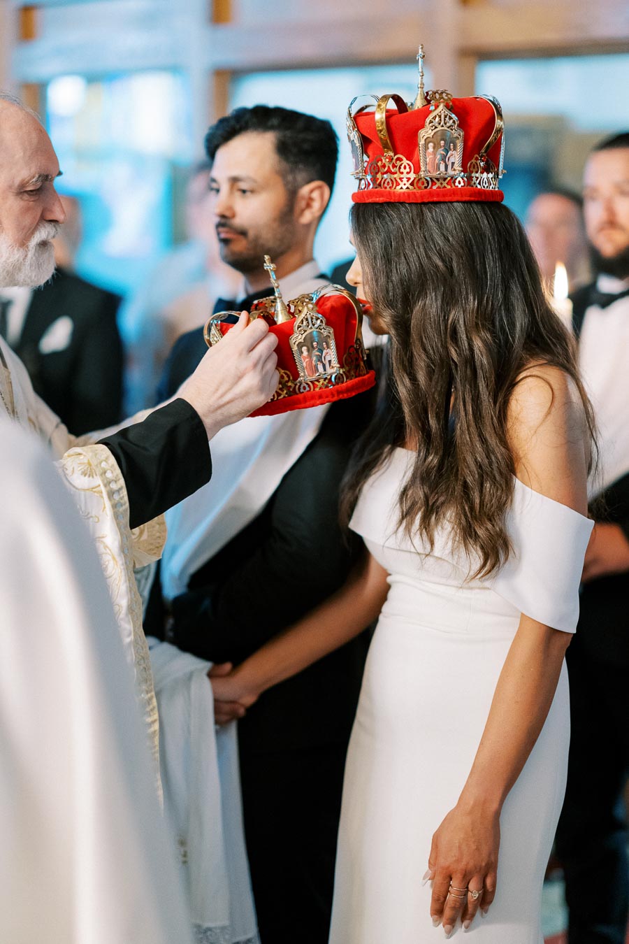 Orthodox wedding ceremony with a bride in a white dress and groom wearing a suit, both adorned with red and gold crowns, during a traditional religious blessing.