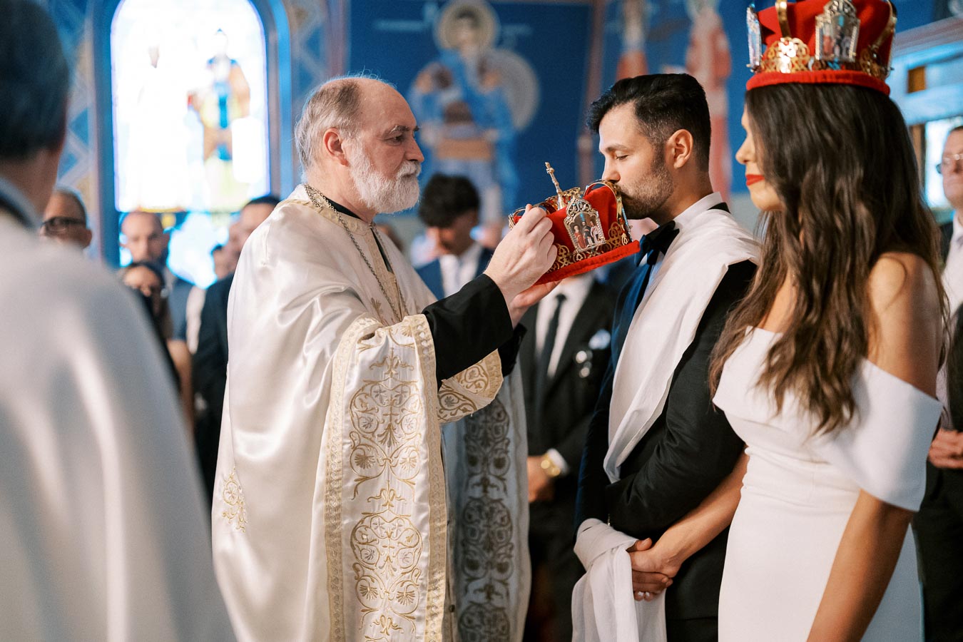 A traditional wedding ceremony in an ornate church, featuring a priest in elaborate robes placing a ceremonial crown on a groom, with the bride standing beside him in a white dress. The background shows religious iconography and stained glass windows.