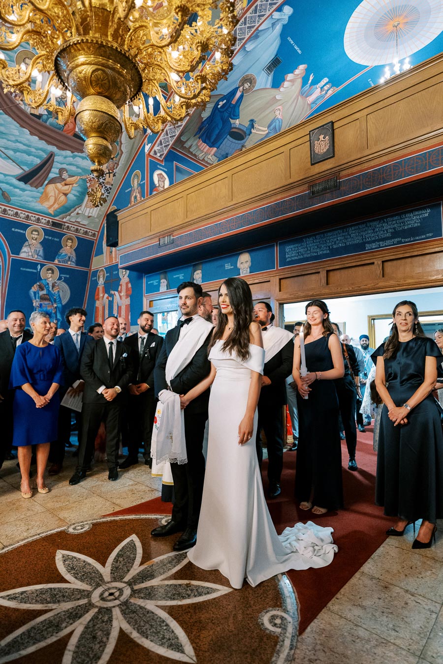 A couple standing at the altar during a traditional church wedding ceremony, surrounded by elegantly dressed guests, with a beautifully painted ceiling and ornate chandelier.