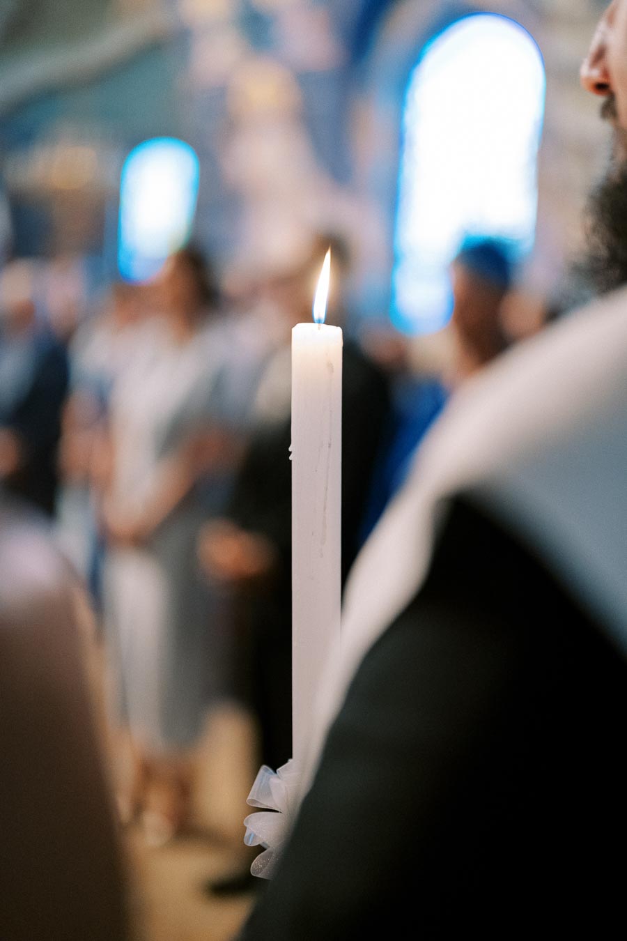 A close-up of a lit white candle in a soft-focus indoor setting with blurred people in the background, suggesting a ceremonial or religious gathering.