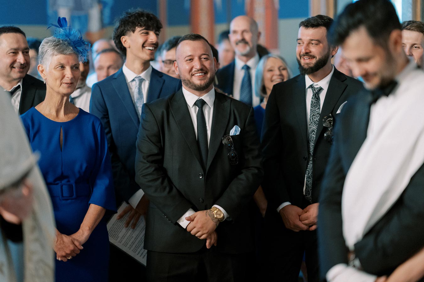 Group of elegantly dressed people smiling and laughing at a formal event, with one woman wearing a blue dress and fascinator.