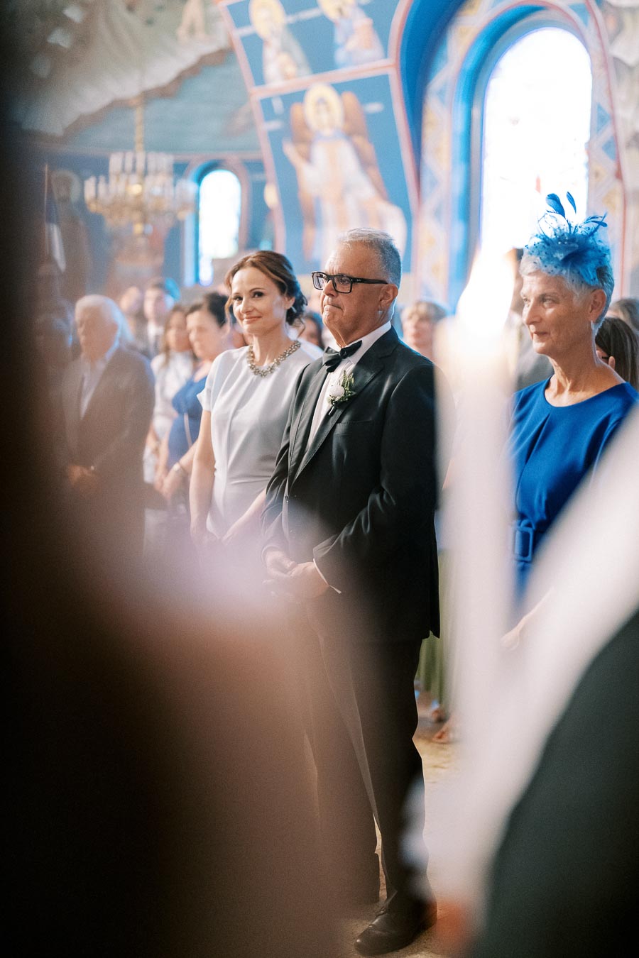 Elderly man in a suit and bowtie stands alongside two women in a church during a ceremony, with vivid blue stained glass and religious murals in the background.