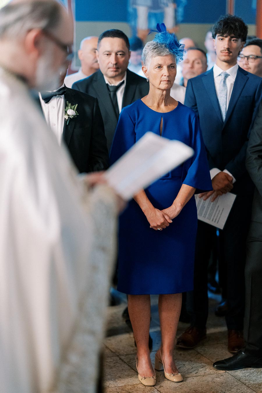 A woman in a royal blue dress and hat stands attentively at a formal event, surrounded by well-dressed attendees in suits and ties, with a focus on a person speaking in the foreground.
