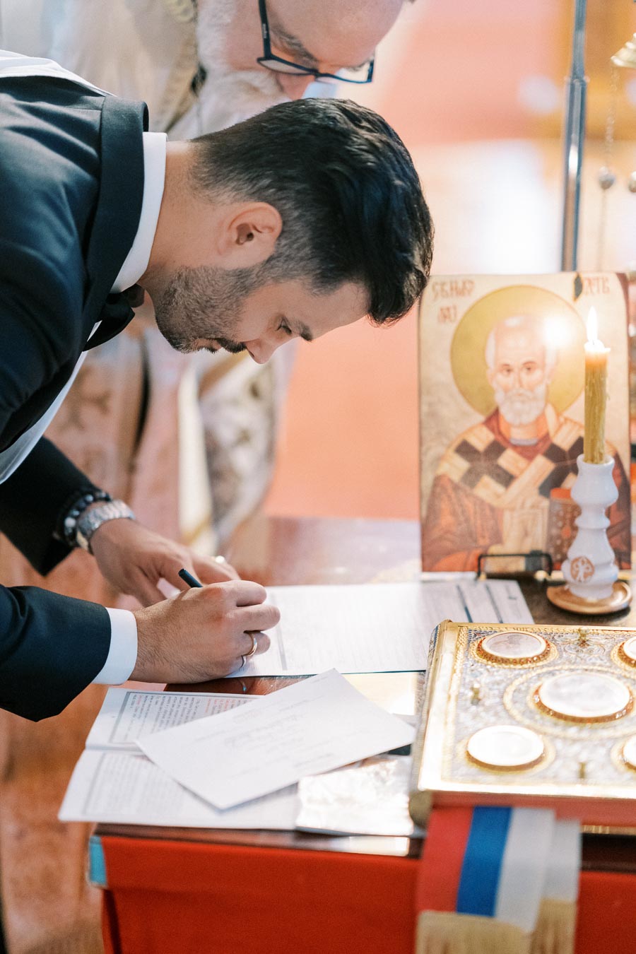 Man in formal attire signing documents on a table during a religious ceremony, with icons and a lit candle beside him.