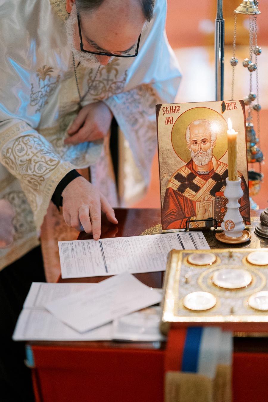 A priest examining documents on a table next to an icon of a saint, accompanied by a lit candle and religious artifacts during a church ceremony.