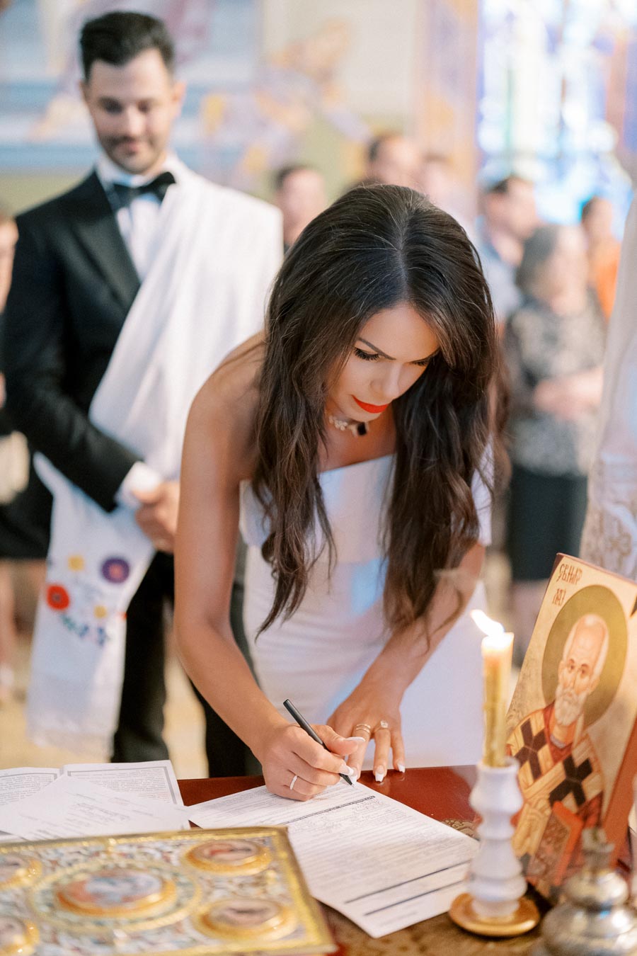 A woman in a white dress signs a document at a ceremonial table adorned with religious icons and a lit candle, with a man in formal attire standing behind her, suggesting a wedding or formal celebration scene.