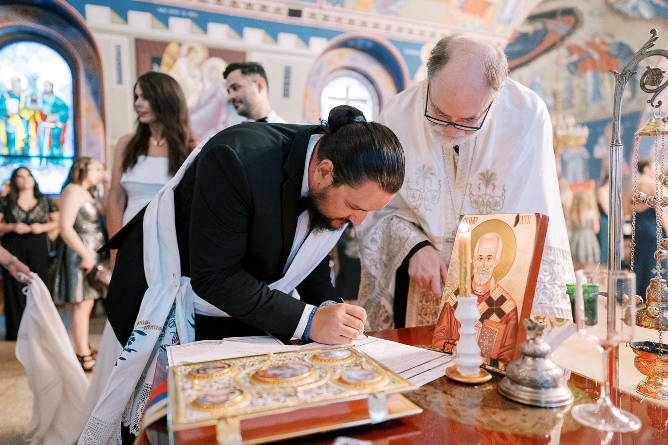 A man signing a document during a traditional ceremony inside a beautifully decorated church, accompanied by a priest in ceremonial robes. The scene includes religious icons and candles, with attendees witnessing the event.