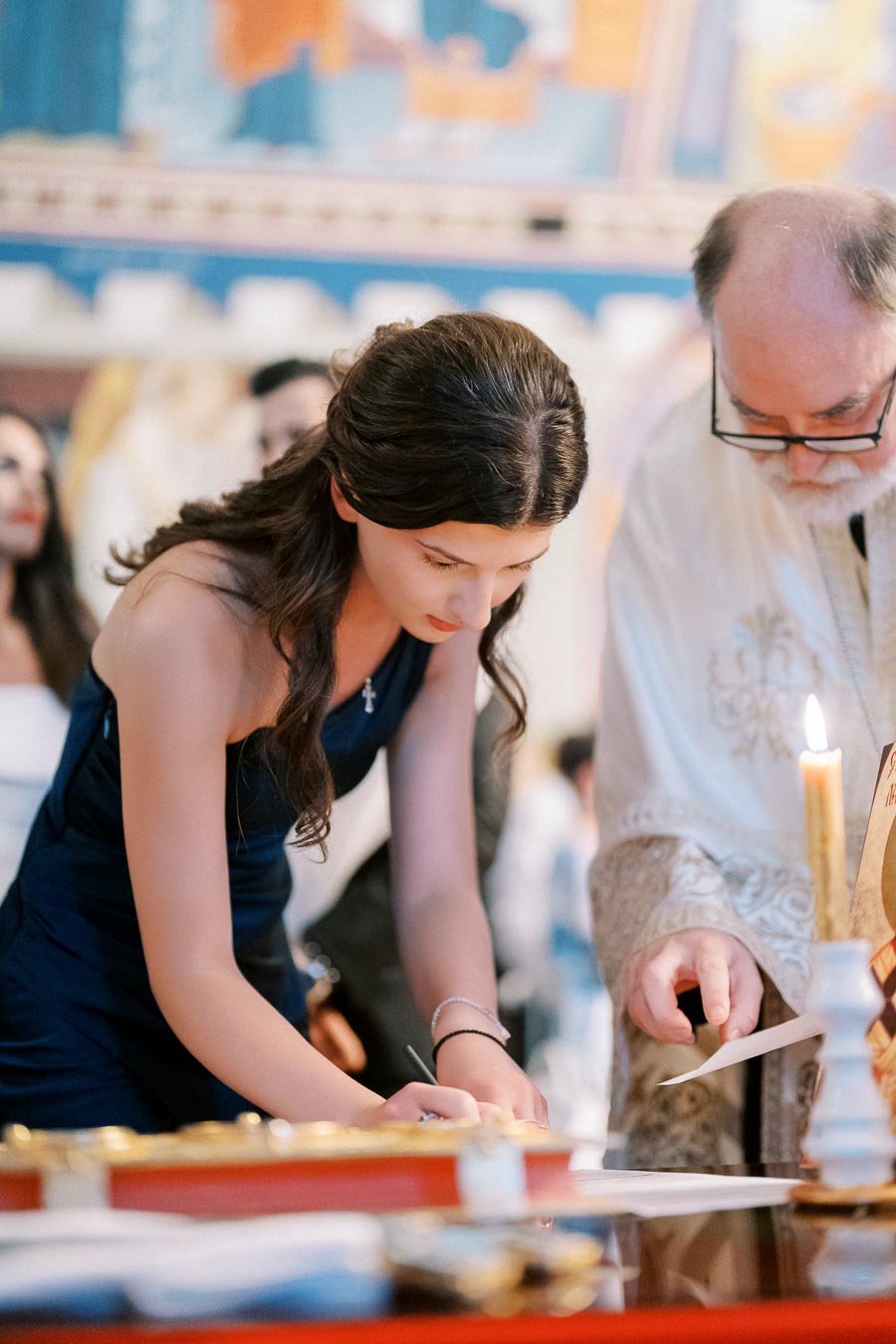 Young woman in a dark blue dress signs a document during a formal ceremony, with a man in traditional attire guiding her.