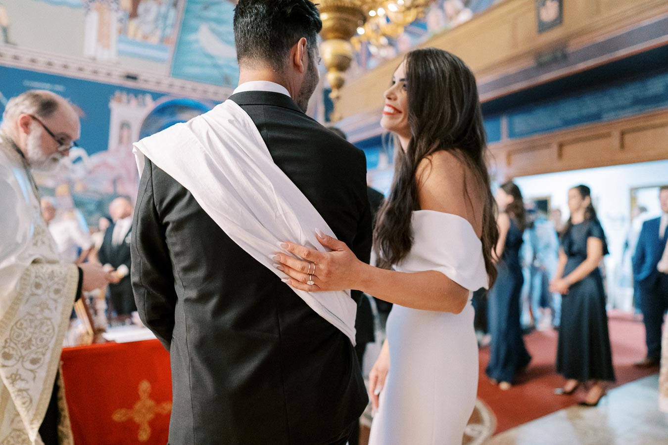 Bride and groom standing together in an ornate church during their wedding ceremony, surrounded by guests and a priest in traditional attire.