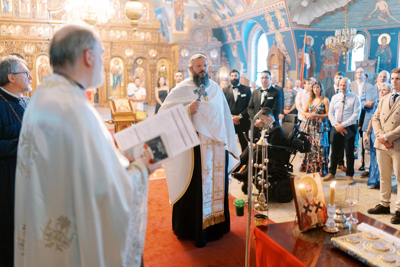 Orthodox church ceremony with priest and congregation gathered inside richly decorated interior, featuring religious icons and ornate architecture.