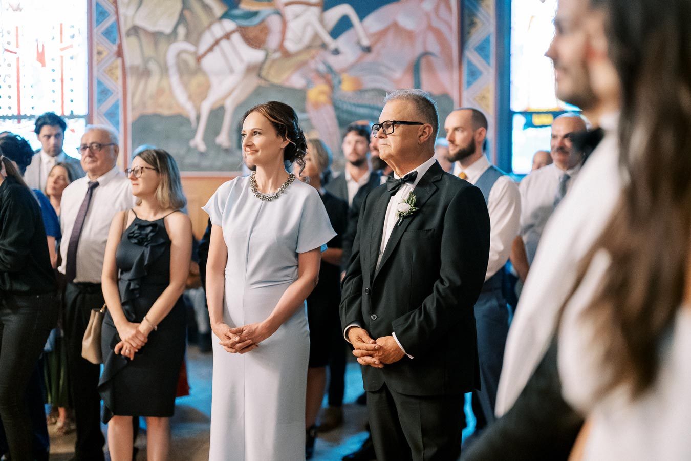 A group of elegantly dressed people stand attentively during a formal event in a decorated room.