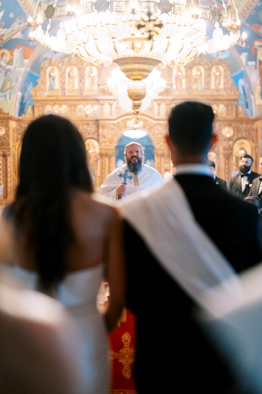 A couple in wedding attire standing before a priest in an ornate church, featuring a grand chandelier and intricate gold decorations.