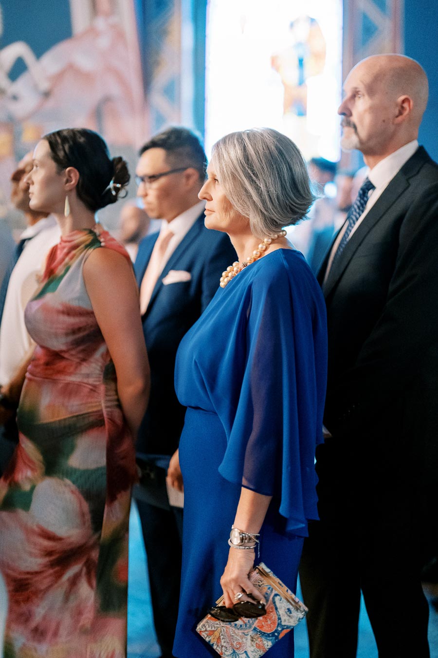 A group of elegantly dressed individuals attending a formal event, featuring a woman in a vibrant blue dress holding a patterned clutch.