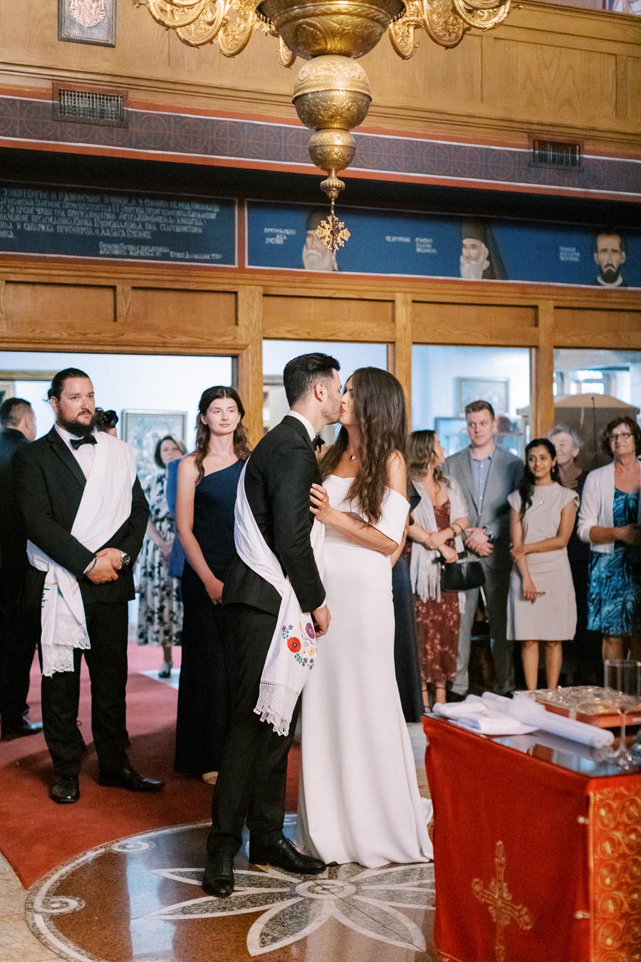 A bride and groom share a kiss during a traditional wedding ceremony in a church, surrounded by well-dressed guests with ornate decorations in the background.