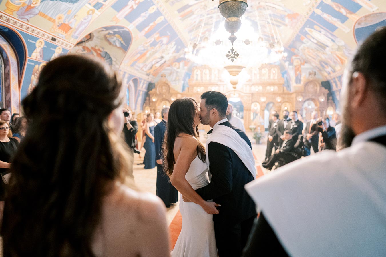 Couple shares a kiss during a traditional wedding ceremony inside an ornately decorated church, surrounded by guests and vibrant frescoes.