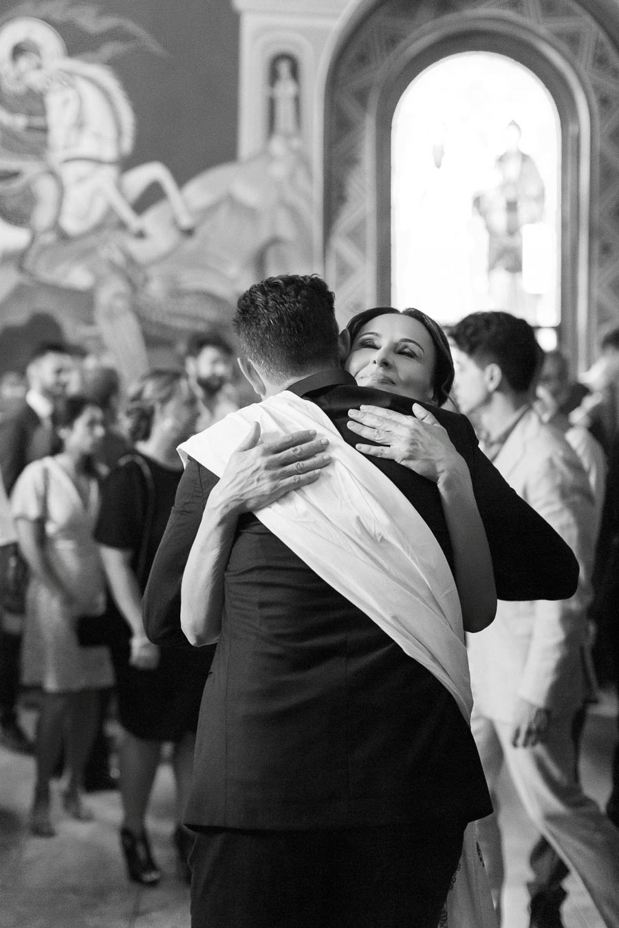A black and white photo of a heartfelt embrace between two people in a crowded setting, with one wearing a ceremonial sash. The background features blurred attendees and ornate architectural details, suggesting a celebratory or formal event.