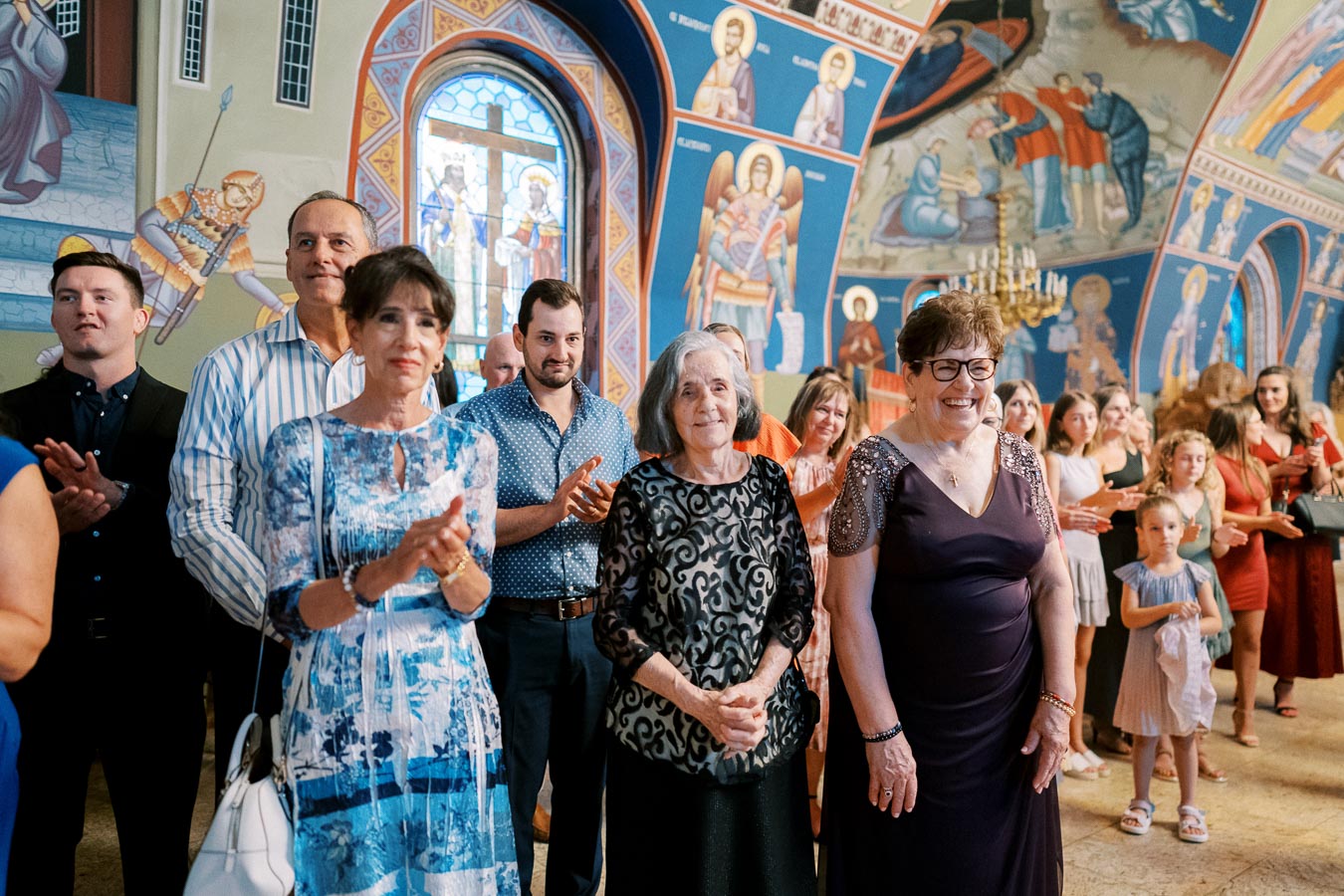 A group of people inside a beautifully decorated church, clapping and smiling during a ceremony. The background features vibrant religious murals and stained glass, creating a joyful and spiritual ambiance.