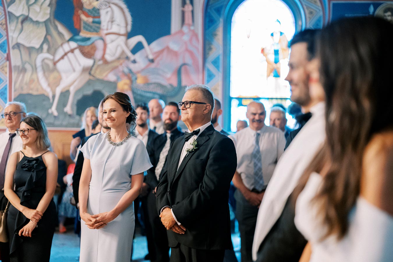 A group of well-dressed guests smiling and standing inside a beautifully decorated venue with vibrant murals and stained glass in the background, capturing a joyful and elegant event.