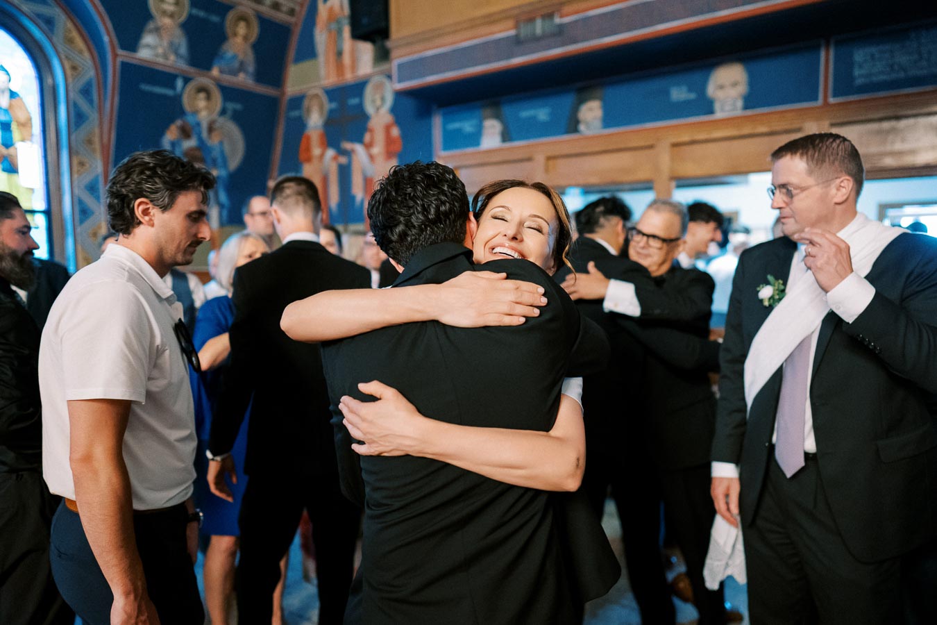 A joyful gathering in a decorated venue, featuring a woman smiling warmly while embracing a man in a suit, surrounded by other guests in formal attire.