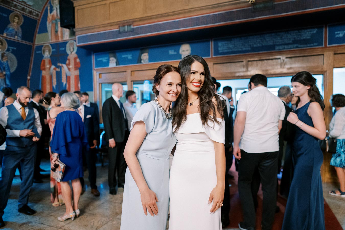 Two women smiling and posing together in elegant dresses at a formal event, surrounded by other guests in a room with ornate wall paintings.