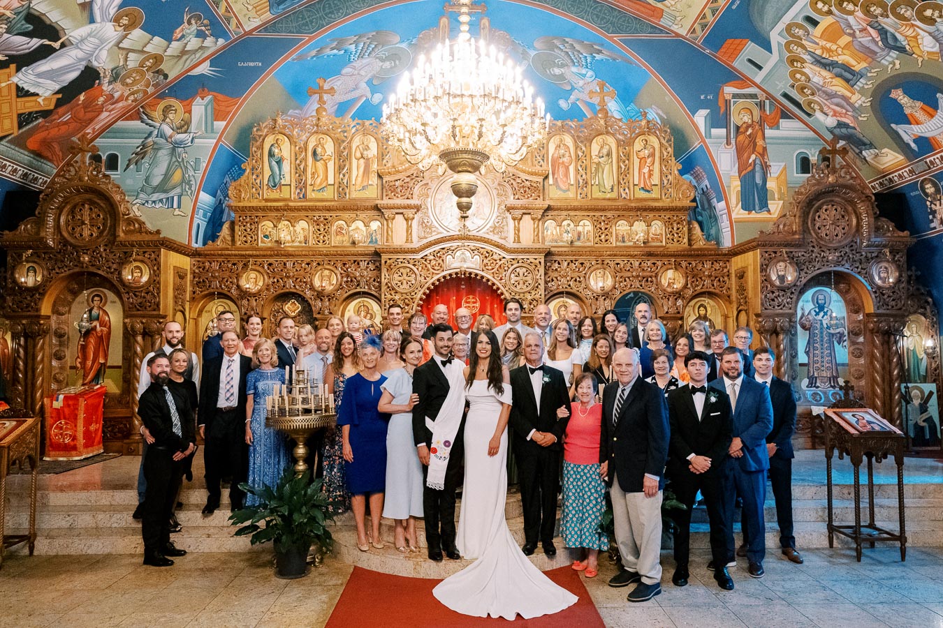 A large group of people posing inside an ornate church with colorful murals and intricate woodwork, gathered for a wedding ceremony. A couple in wedding attire stands at the center under a grand chandelier.