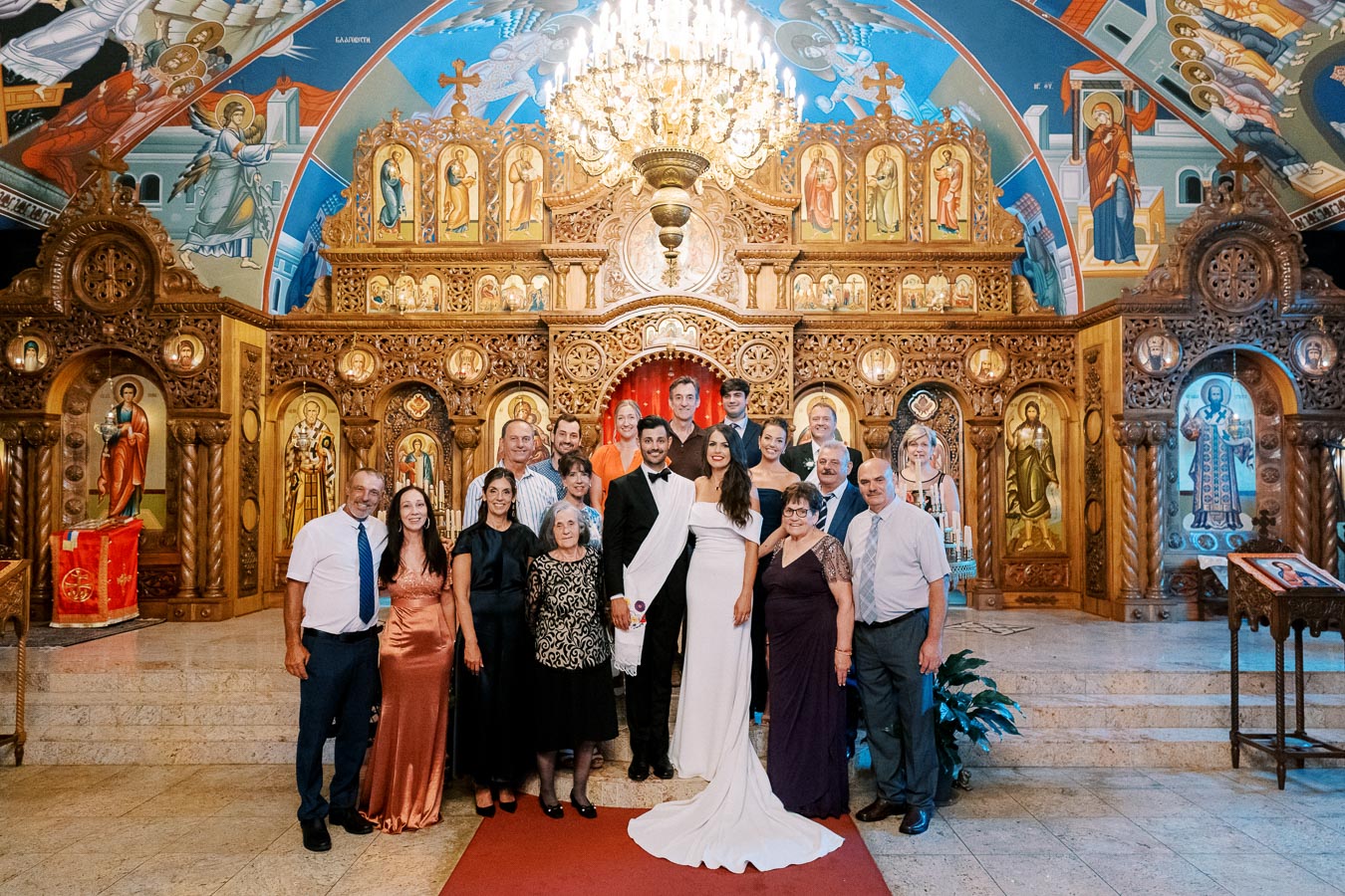 A group of people posing inside a beautifully decorated church with ornate wooden carvings and religious icons, under a large chandelier.