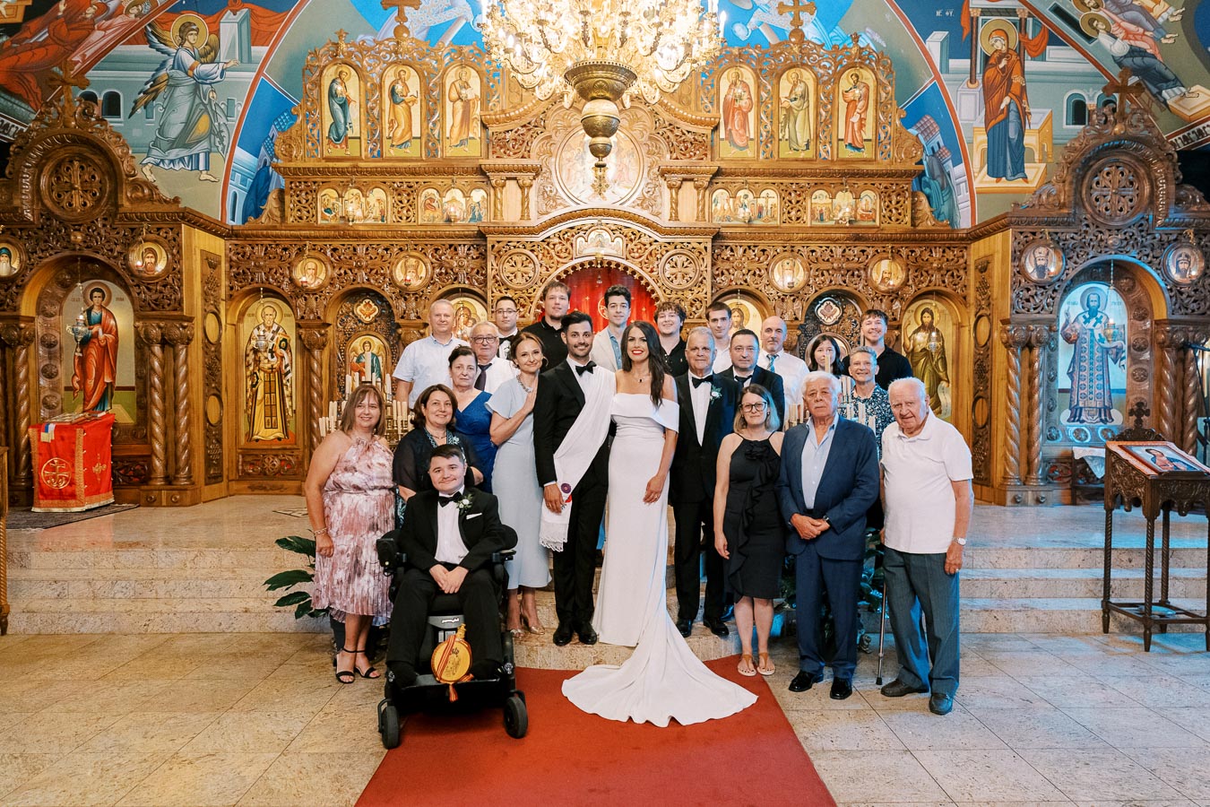 A wedding party poses in an ornately decorated church with wooden carvings and religious icons; the bride and groom stand in front surrounded by family and friends.