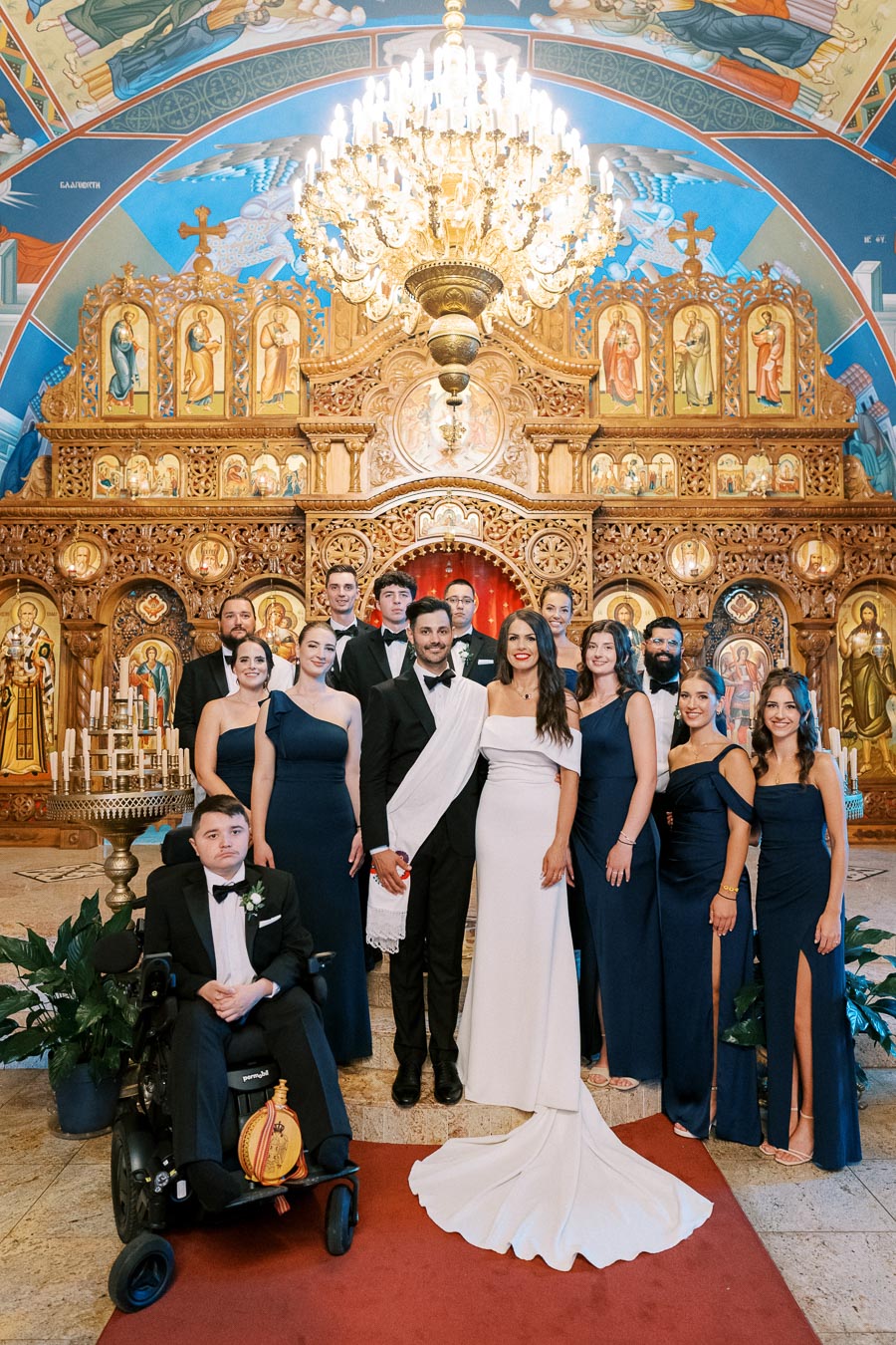A wedding party poses in elegant attire at an ornately decorated church with a grand chandelier and richly detailed iconography in the background. The bride wears a stunning white gown while bridesmaids are in navy dresses.