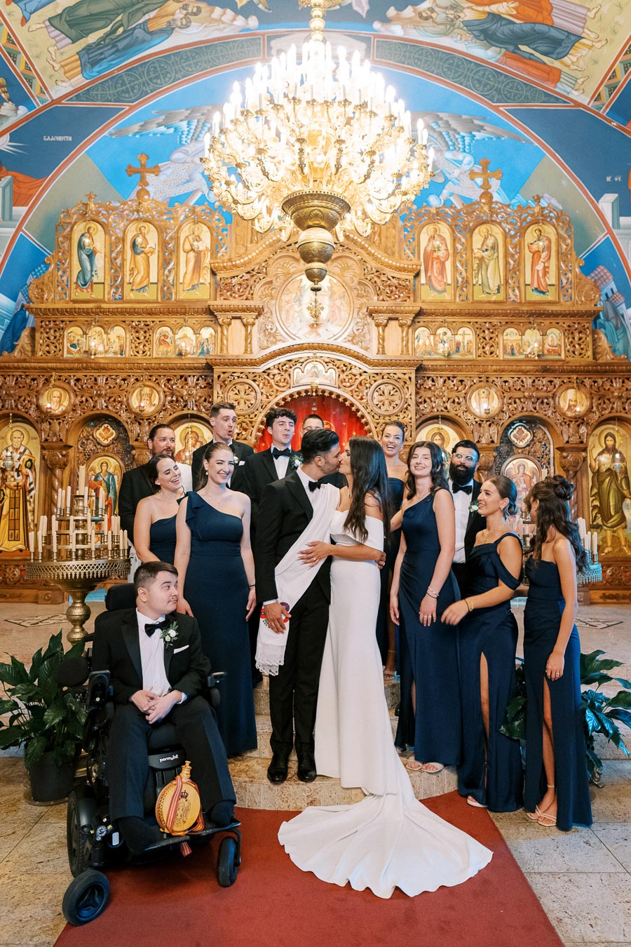 Bride and groom kissing in a beautifully decorated Orthodox church surrounded by a joyful wedding party wearing elegant formal attire.