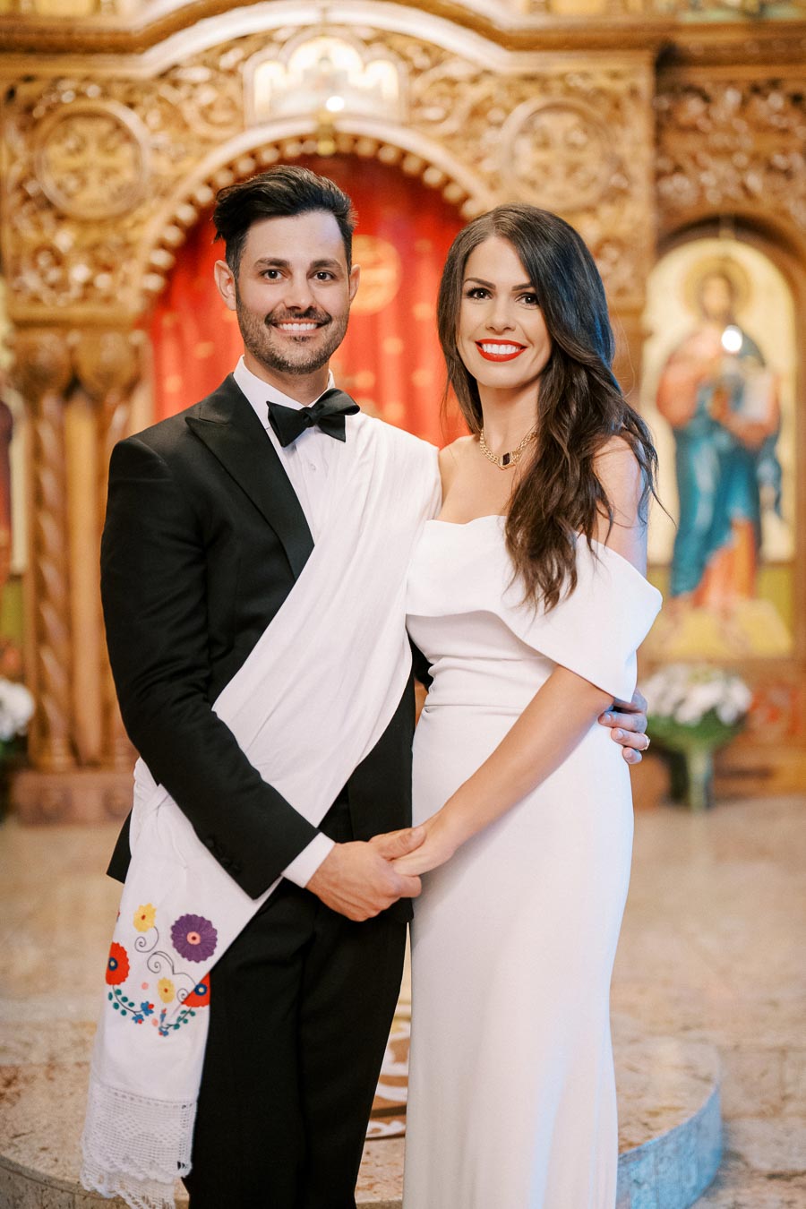 A couple dressed in elegant formal attire stands together in a beautifully decorated church interior, with intricate wooden carvings and religious artwork in the background, possibly celebrating a special occasion.