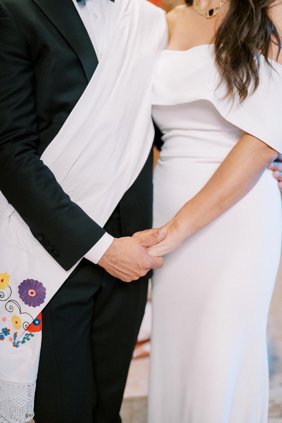 A couple holding hands during a wedding, wearing elegant attire. The groom is in a black suit with a white shawl featuring colorful floral embroidery, while the bride is in an off-shoulder white dress.