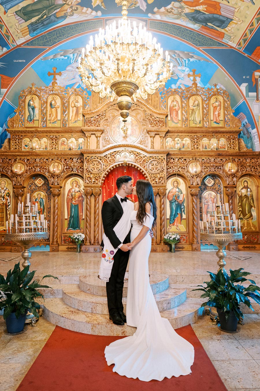 A bride and groom in elegant attire standing in front of an ornate altar inside a beautifully decorated church, beneath a grand chandelier, showcasing a traditional and culturally rich wedding setting.