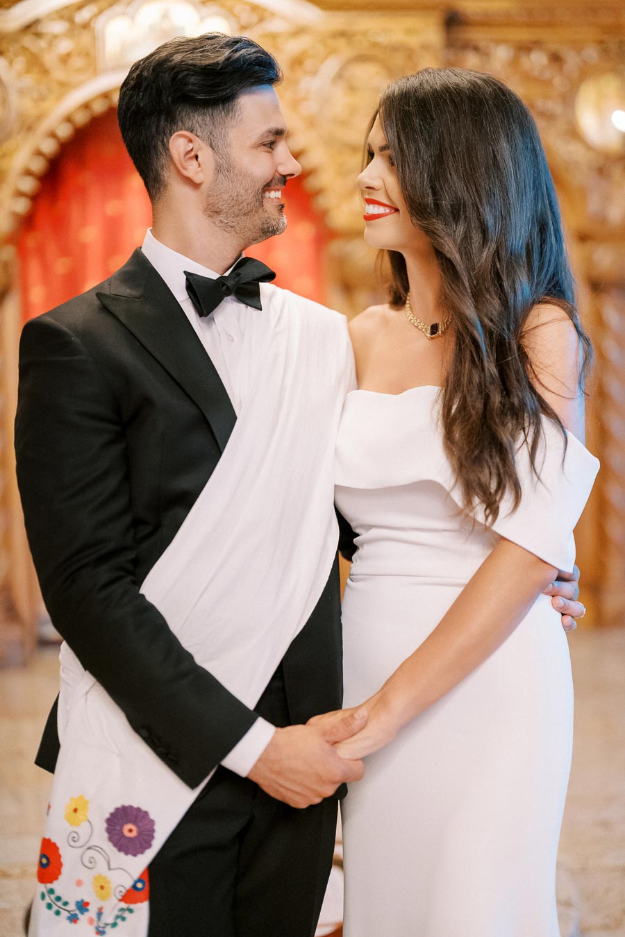 A couple dressed in elegant, formal attire, smiling at each other in a beautifully decorated indoor setting. The man is wearing a black suit with a bow tie and a white sash with colorful floral embroidery, while the woman is in a white off-shoulder gown.