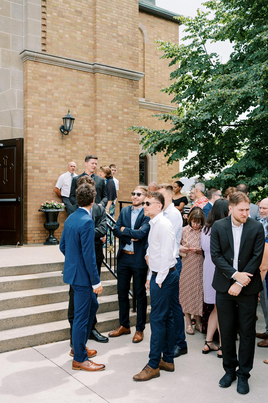Group of people in formal attire gathered outside a brick building, possibly a church or event venue, engaged in conversation beside a set of stairs and a large tree.