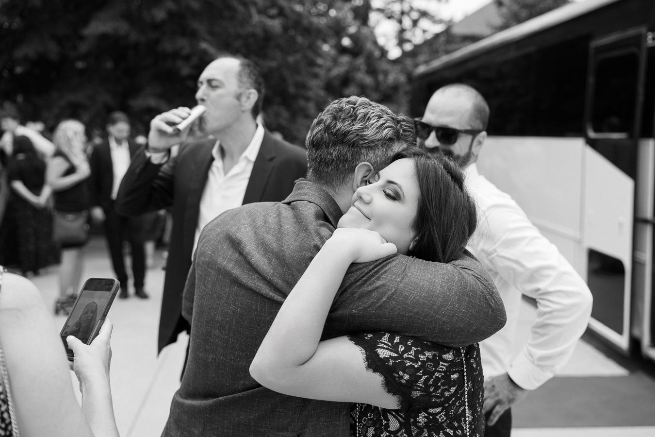 A black and white photo capturing a heartfelt embrace between a man and a woman at an outdoor event, with onlookers in the background and a bus parked nearby.