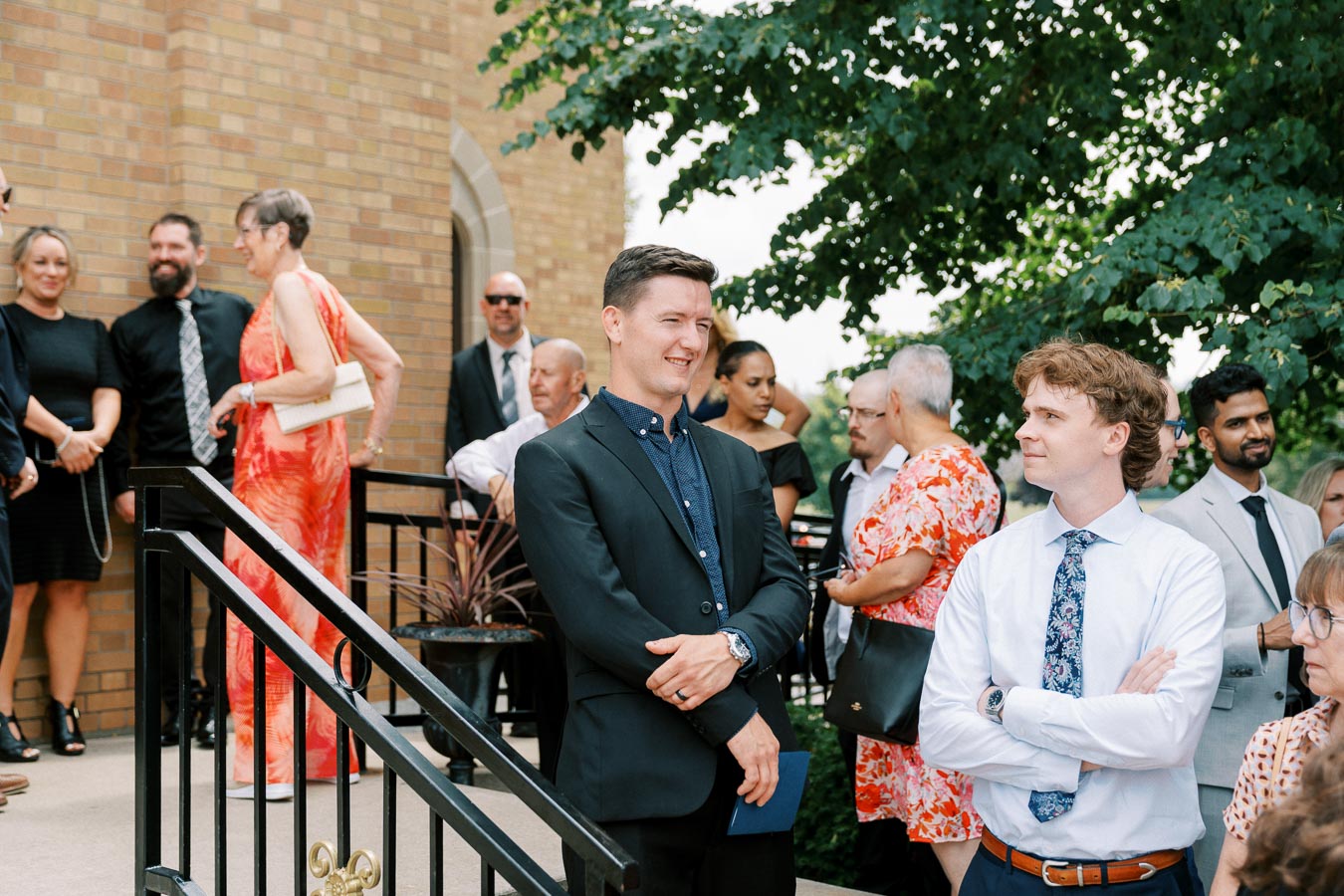 A group of well-dressed people socializing outside a building, with men in suits and women in dresses, surrounded by greenery.