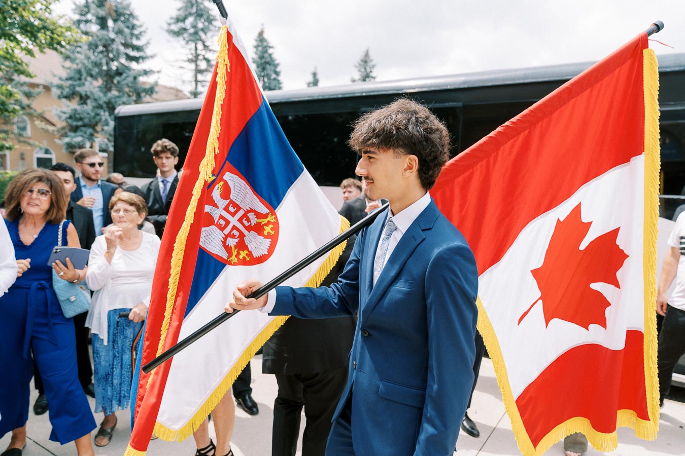Young man in a suit holding Serbian and Canadian flags during an outdoor gathering, surrounded by a group of people in formal attire.