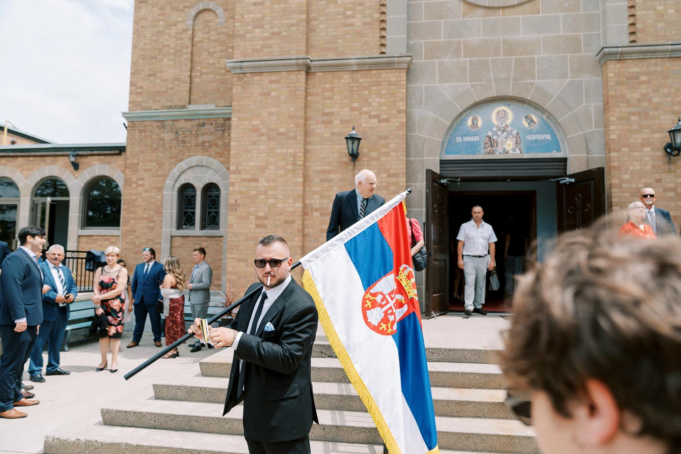 A group of people, dressed in formal attire, stand outside a brick building with arched windows. In the foreground, a man in a suit holds the Serbian flag, while others converse near the entrance adorned with religious art.