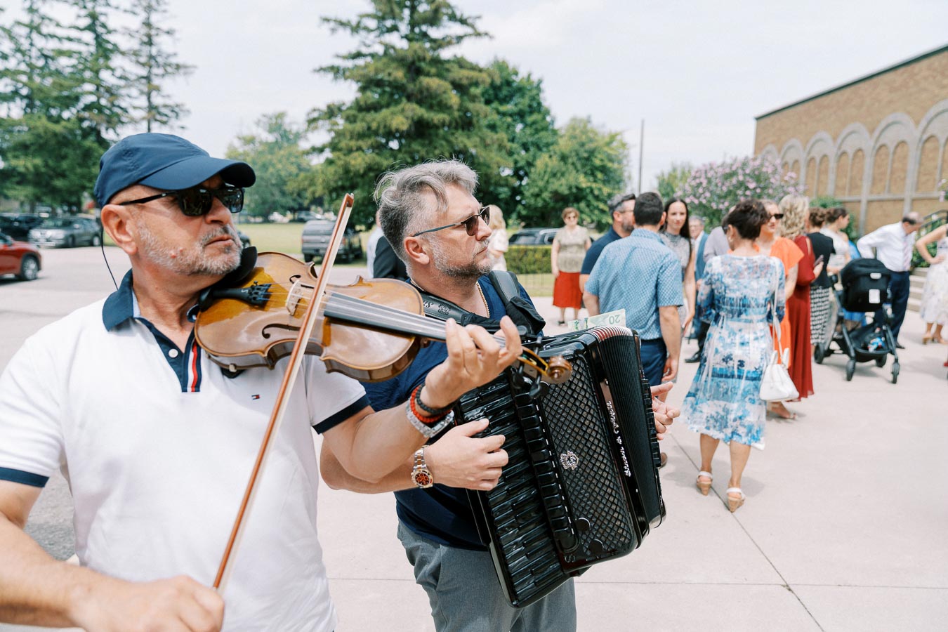 Two musicians playing violin and accordion at an outdoor event, surrounded by a crowd in summer attire.