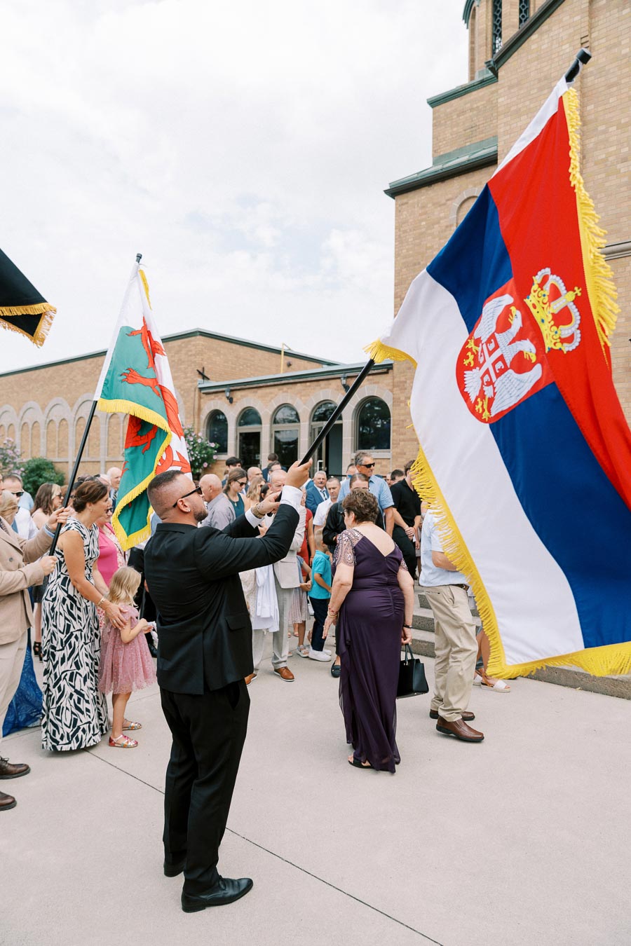 A man in a black suit proudly waves a Serbian flag next to a crowd gathered outside a church, celebrating a cultural event.