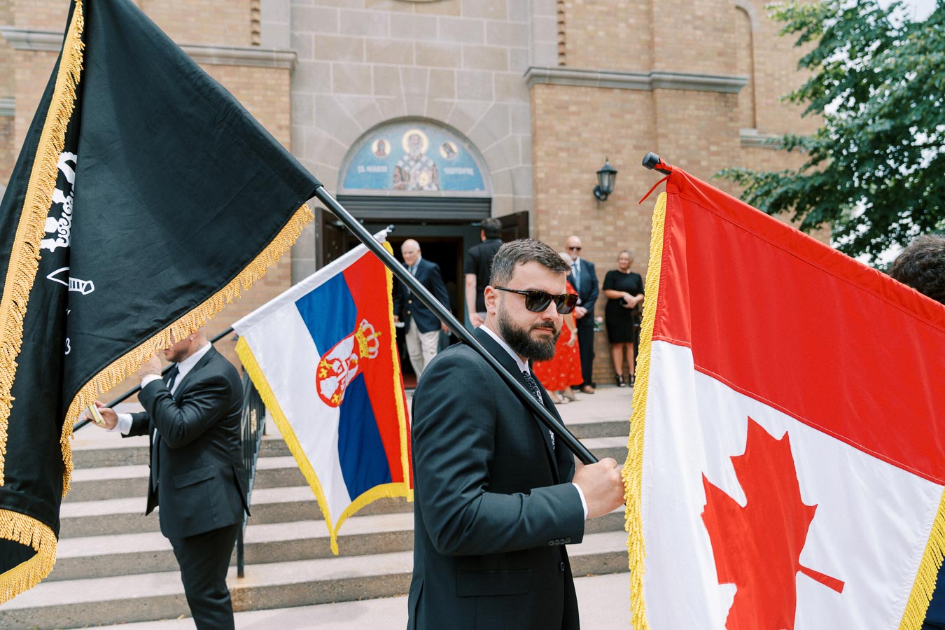 A man in a suit holding a Canadian flag with others holding various flags in front of a church entrance.