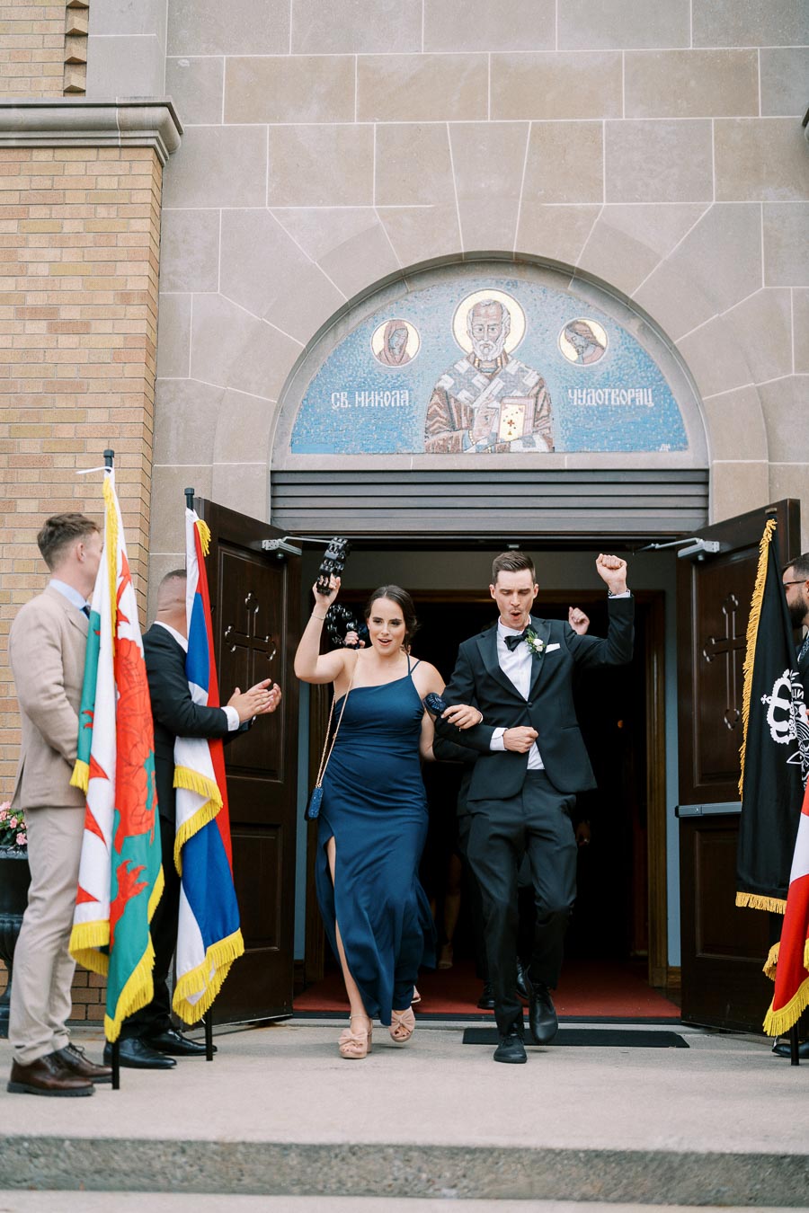 A couple exits a church celebrating, with friends cheering them on amidst colorful flags. The historic building features a mosaic above the entrance.