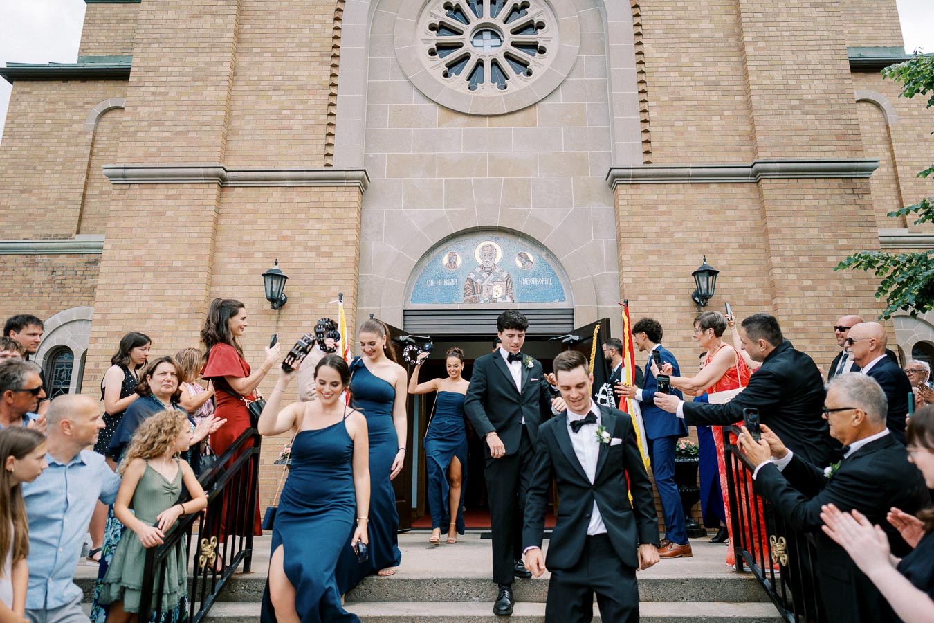 Wedding party exiting a church, with bridesmaids in blue dresses and groomsmen in tuxedos, surrounded by smiling guests capturing the moment on their phones.