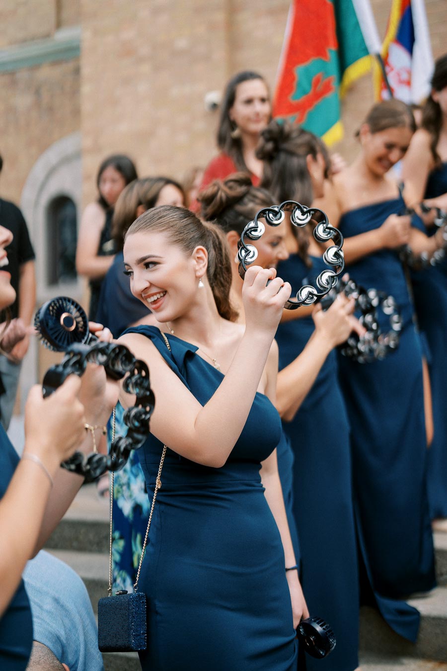 A group of women in elegant blue dresses joyfully playing tambourines outside a building, with flags in the background, creating a festive and celebratory atmosphere.