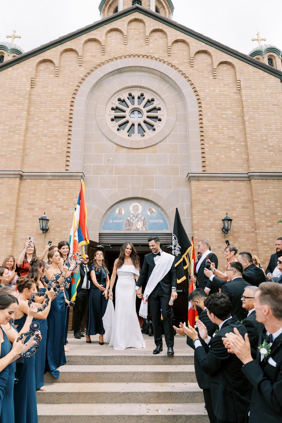 Newlyweds exiting church amid celebration, with guests clapping and holding tambourines, surrounded by a historic brick church facade and religious icons.