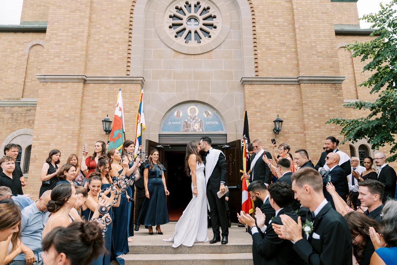 A happy couple shares a kiss on the steps of a church after their wedding ceremony, surrounded by cheering friends and family dressed in formal attire, holding festive decorations.