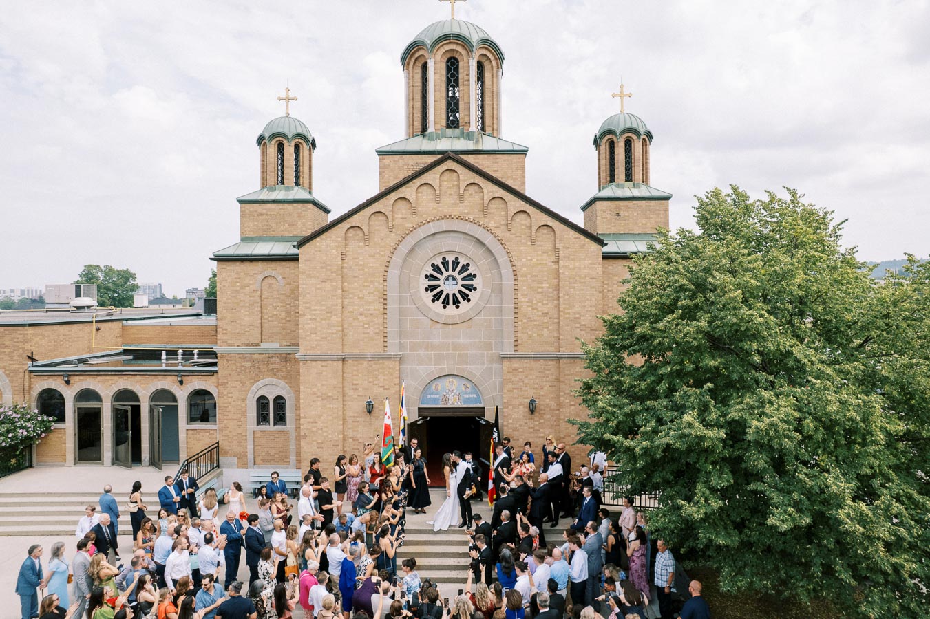 Wedding celebration outside a large brick Orthodox church with a crowd of guests gathered around, featuring three green-roofed towers topped with crosses and lush trees in the foreground.