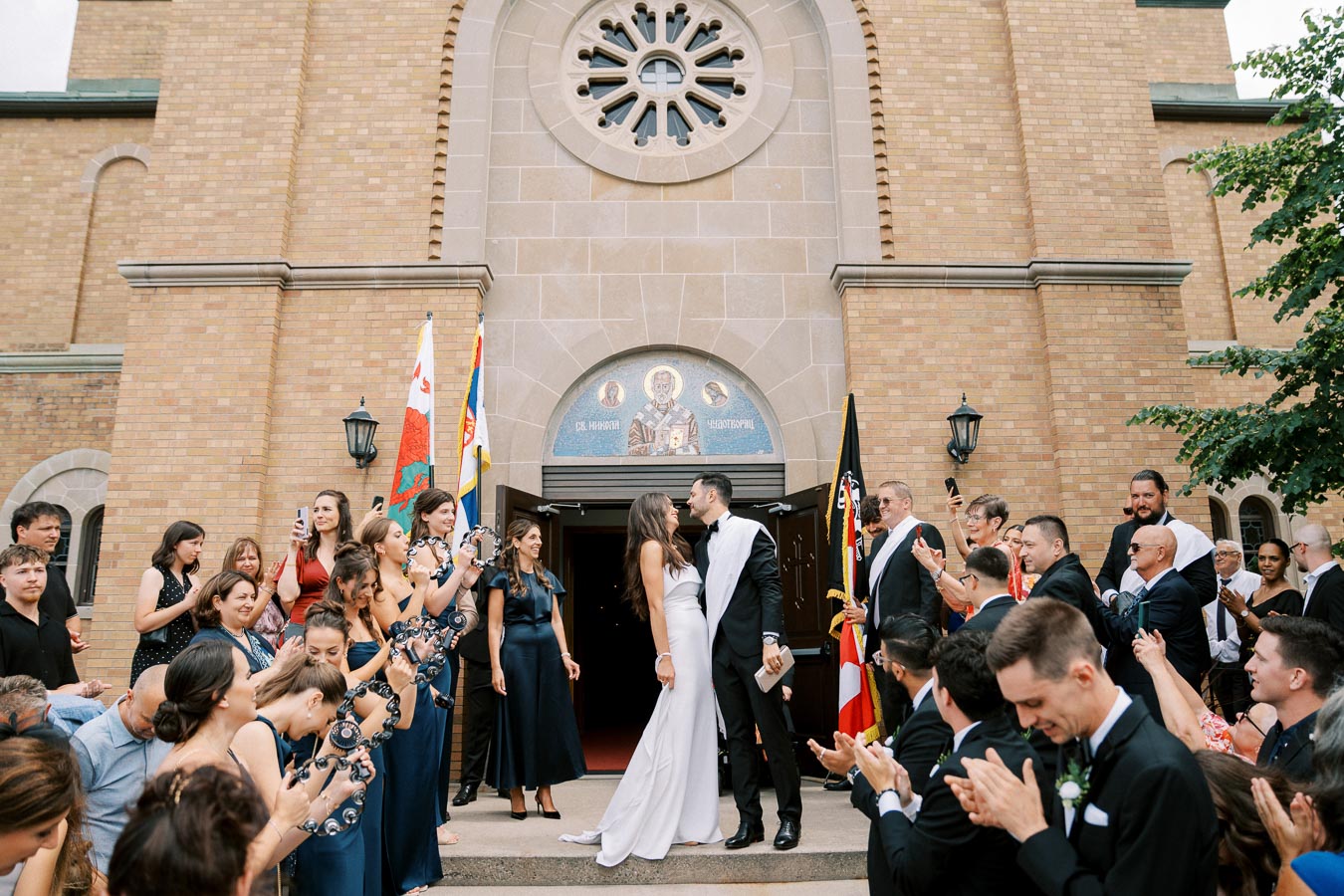 A newlywed couple shares a kiss in front of a church, surrounded by cheering wedding guests. The bride and groom stand at the entrance, adorned with flags and religious symbols. Joyful friends and family, dressed in formal attire, clap and capture the moment.